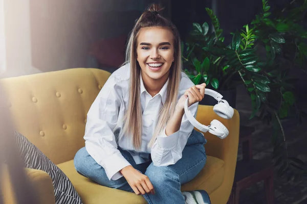 Woman sits on yellow sofa and holds in her hand white wireless headphones. Millennial girl looks the camera with smile — Stock Photo, Image