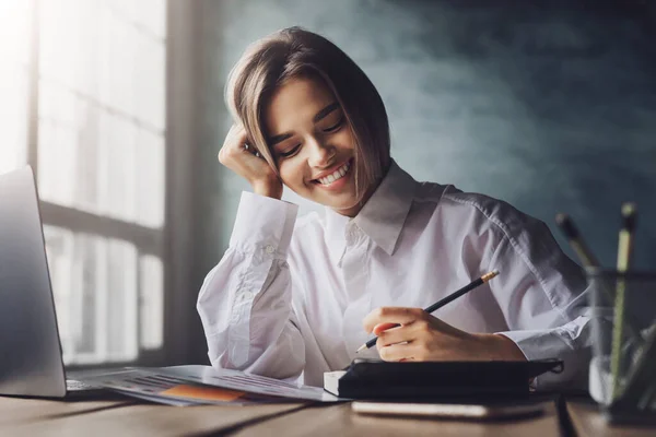 Chica sonriente sentada en el escritorio, tomando notas con bolígrafo en el cuaderno. Estudiante femenina estudiando delante de la laptop — Foto de Stock