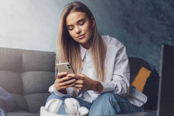 Pretty casually dressed girl with long hair sitting on coach and texting on smartphone. Young lady using portable mobile — Stock Photo, Image