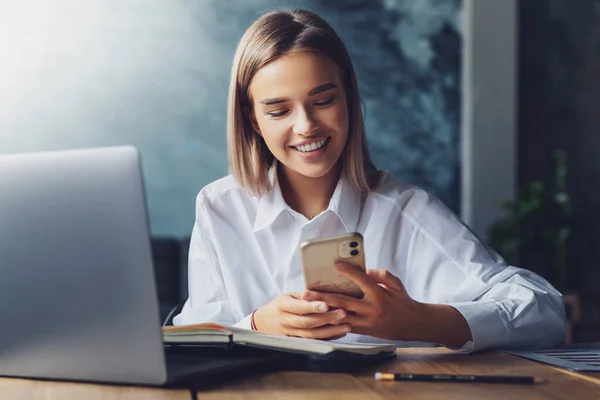 Pretty girl smiling and texting on smartphone. Cheerful female having break at workplace, communicating with friends