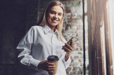 Gülümseyen kız elinde akıllı telefon ve bir fincan kahve tutuyor. Kızlar iş için cep telefonu kullanır