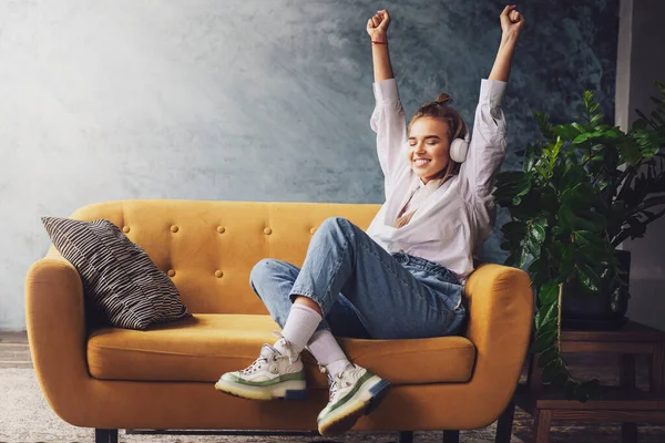 Chica en camisa blanca se sienta en el sofá y disfruta de la transmisión de audio, podcasts. Joven mujer milenaria disfruta de tiempo libre en casa . —  Fotos de Stock