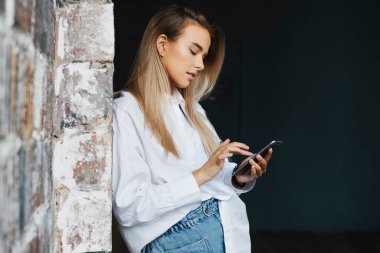 Beyaz gömlekli güzel bir iş kadını ofiste tuğla duvarın yanında duruyor, akıllı telefonu elinde tutuyor..