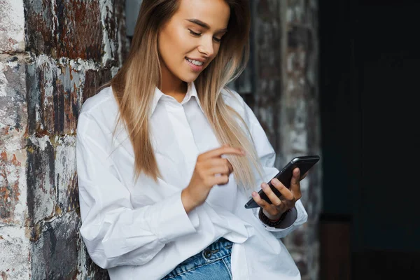 オフィスのレンガの壁の近くに白いシャツを着た美しい若いビジネス女性は、彼女の手にスマートフォンを保持します. — ストック写真