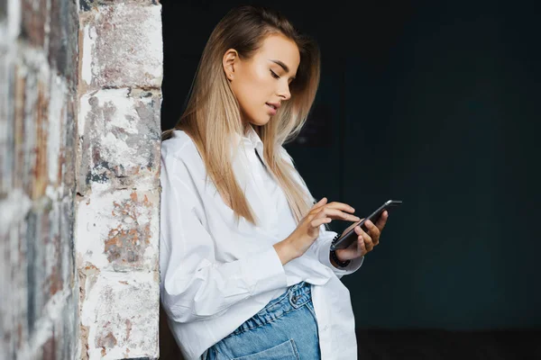 オフィスのレンガの壁の近くに白いシャツを着た美しい若いビジネス女性は、彼女の手にスマートフォンを保持します. — ストック写真