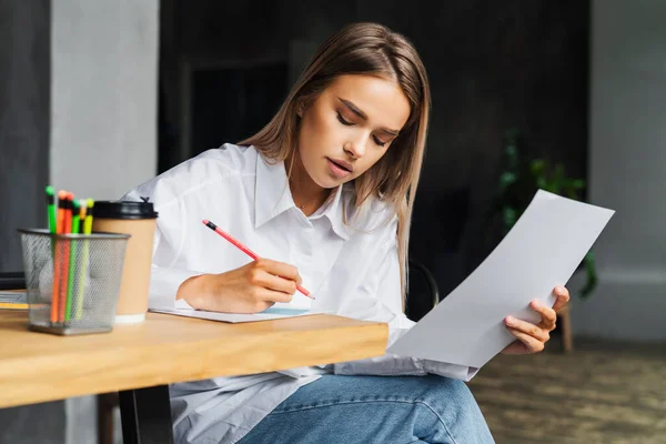 Hermosa mujer de negocios que trabaja en el proyecto mientras está sentada en la mesa de madera, hace notas en la hoja de papel blanco . — Foto de Stock