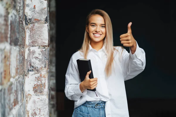 Beautiful blonde girl is standing in white shirt on dark background near brick wall in office, shows approving gesture. — Stock Photo, Image