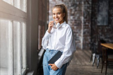 Güzel sarışın kız defterini elinde tutuyor. Beyaz gömlekli kız gülümsüyor ve pencerenin yanında duruyor..