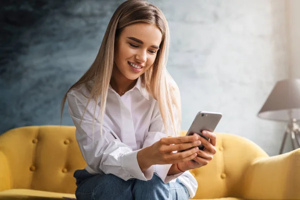 Una bella donna lavora a casa seduta su un divano giallo, usando uno smartphone, scrivendo messaggi, condividendo foto — Foto Stock