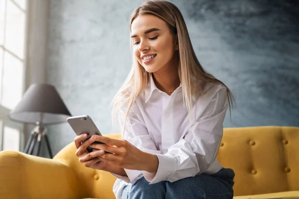 Una bella donna lavora a casa seduta su un divano giallo, usando uno smartphone, scrivendo messaggi, condividendo foto — Foto Stock