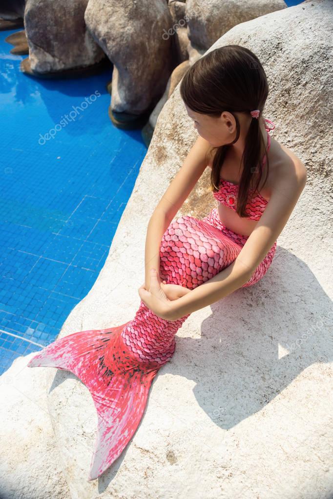 Mermaid girl with pink tail on rock at poolside