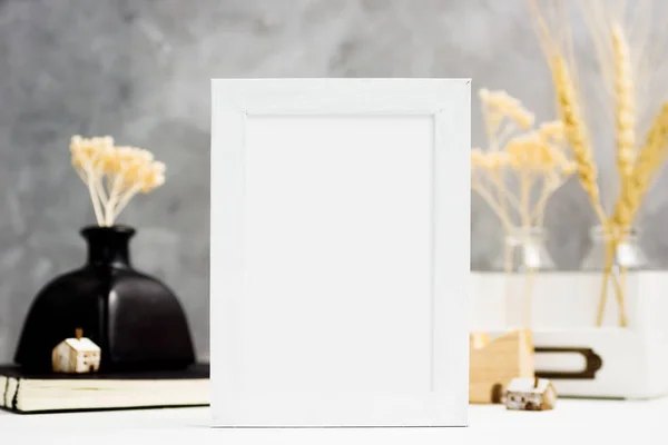 Vertical white Photo frame mock up with dry plants in vase, notebook and wooden houses on shelf. Scandinavian style. Text space