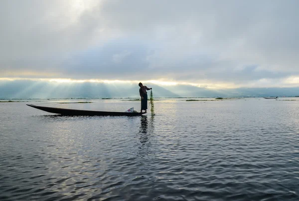 Pescador em barco de madeira lançando uma rede para a captura de peixes no lago no início da manhã antes do nascer do sol — Fotografia de Stock
