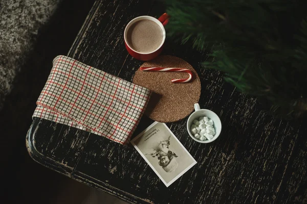 Cocoa in a red cup near a checkered cloth, cards, candy on a stand of cork, cup with marshmallow