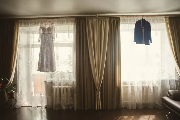 Robe de mariée blanche à une fenêtre, et le costume du marié sur le rack à l'autre boîte avec des rideaux beiges et dentelle mariage en tulle par une journée ensoleillée Images De Stock Libres De Droits