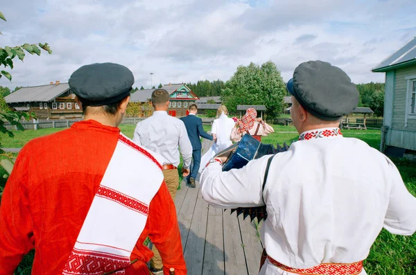 Bruiloft in het Russische dorp. De accordeon in een wit overhemd, folk, een man in een rood shirt en een cap met een witte handdoek op zijn schouder naar de houten huizen op de achtergrond op een houten wandelpad, uitzicht vanaf de achterkant — Stockfoto