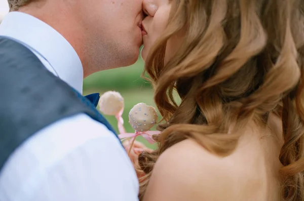 La novia y el novio en un traje azul y pajarita están sosteniendo pastel-pop, besándose en el verano en la naturaleza de cerca —  Fotos de Stock