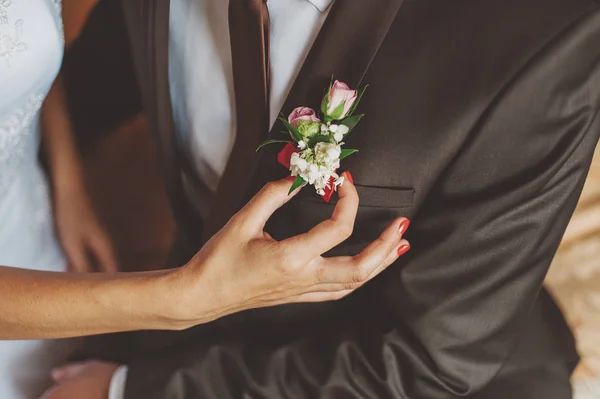 A noiva com a manicura vermelha corrige o boutonniere do pequeno noivo de rosa de rosa em um terno e gravata — Fotografia de Stock