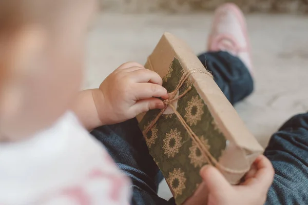Niño pequeño en jeans sosteniendo un regalo en papel kraft con decoración verde y cordel en el estudio en una alfombra blanca, primer plano de las manos —  Fotos de Stock