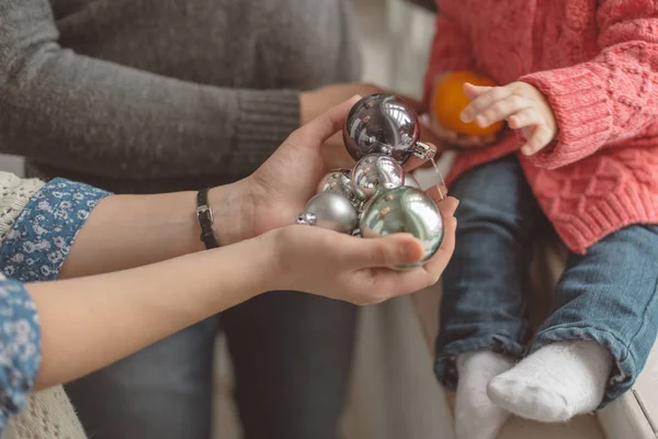 Femme tenant une boule de Noël colorée (grise, argentée, verte), montrant leur enfant en chemisier rose, jeans, famille, studio, gros plan des mains Image En Vente