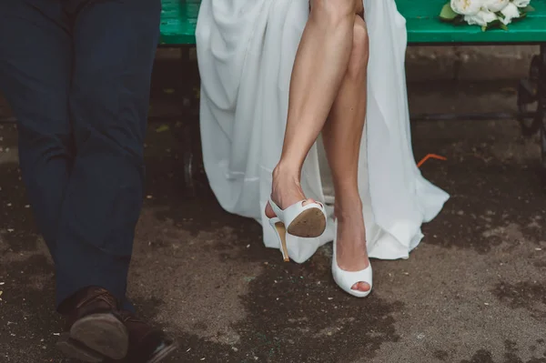 Стройные ноги невесты в белых туфлях с открытым носом и женихом в парке на скамейке летом — стоковое фото