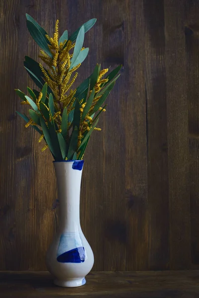 Bouquet de branches jaunes d'acacia, mimosa dans un vase blanc avec des carrés bleus, avec un cou étroit sur le vieux fond brun en bois Photo De Stock