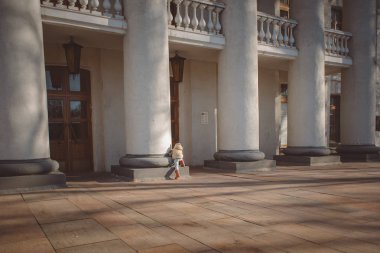 Çocuk bej ceketi giyen, lastik çizme ve şapka erken Bahar güneşli bir tiyatro sütunlardaki yürür