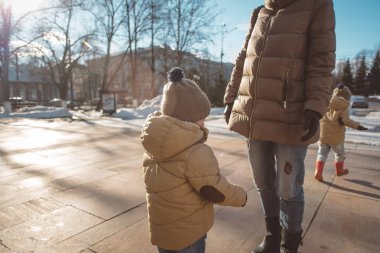 bir bej dutoy ceket, kot pantolon ve bir şapka ile bir ponpon kızı annesi road yakınındaki şehir ile erken Bahar güneşli bir günde, sokaklarda kar yürür