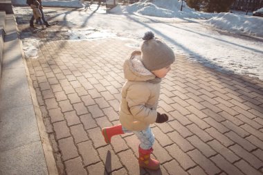 küçük kız bej ceket, kot ve erken Bahar güneşli bir günde, bir parkta çalışan pembe lastik çizme eriyen kar ve mavi gökyüzü, Bina