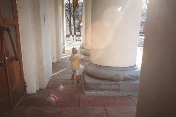 Маленька дівчинка в бежевій куртці, джинсах і гумових чоботях біжить навколо, граючи навколо колон біля входу в театр, дерев'яні двері, ранній весняний сонячний день — стокове фото
