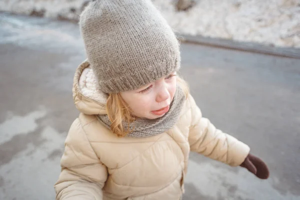 Petite fille blonde dans un chapeau gris tricoté avec pompon et une veste beige pleurer sur la route au début du printemps, fin de l'hiver Images De Stock Libres De Droits