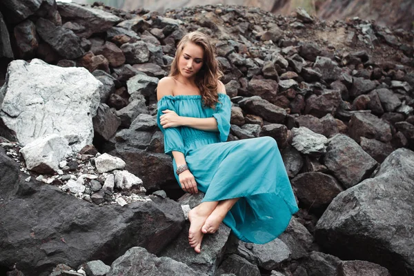 Vackra drömmande blond flicka i en ljus turkos klänning sitter på klipporna — Stockfoto
