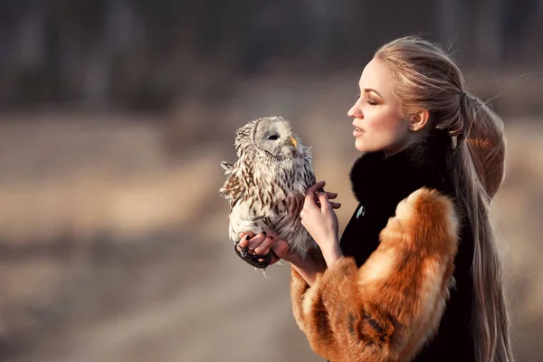 Belle femme en manteau de fourrure avec un hibou sur le bras. Blonde aux cheveux longs dans la nature tenant un hibou. Image romantique délicate d'une fille — Photo