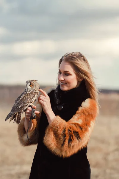 En vakker kvinne i pelskåpe med en ugle på armen. Blond med langt hår i naturen som holder en ugle. Romantisk, ømtålig bilde av en jente – stockfoto