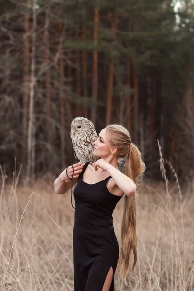 En vakker kvinne i svart kjole med en ugle på armen. Blond med langt hår i naturen som holder en ugle. Romantisk, ømtålig bilde av en jente – stockfoto