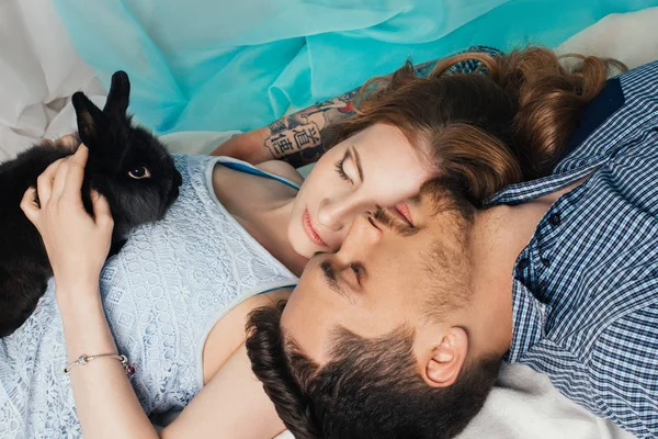 Casal amoroso deitado perto da janela com um coelho nas mãos. Sentimentos ternos e românticos dos recém-casados. Mulher bonita com cabelos longos e um homem com tatuagens em suas mãos — Fotografia de Stock