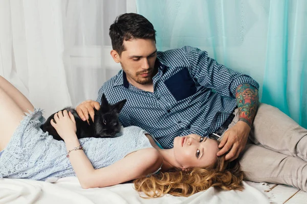 Aimant couple couché près de la fenêtre avec un lapin sur les mains. Sentiments tendres et romantiques des jeunes mariés. Belle femme aux cheveux longs et un homme avec des tatouages sur les mains — Photo