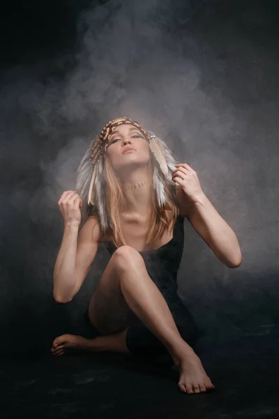 Mujer perfecta vestida de indios americanos en humo sobre un fondo gris. Un sombrero hecho de plumas. Misteriosa manera mística, cuerpo sexy, hermosa espalda. Rubia atractiva con una cara hermosa —  Fotos de Stock