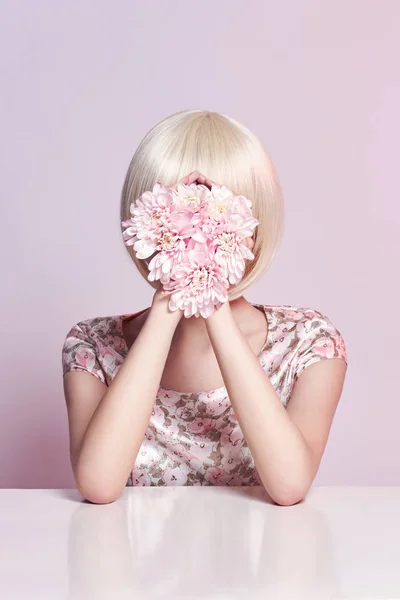 Moda mulher retrato de arte em vestido de verão e flores em sua mão com uma maquiagem contrastante brilhante. Criativa beleza foto meninas sentadas à mesa em um fundo rosa contrastante com sombras coloridas . — Fotografia de Stock