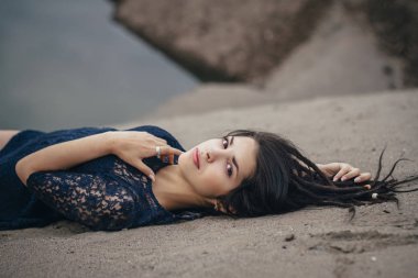 Göle kuma bulutlu bir günde yalan arka plan üzerinde bir kadın esmer portre yaşam tarzı. Romantik, yumuşak, mistik, dalgın bir kız görüntüsü. Kız oryantal görünüm, kum üzerinde yalan rüyalar