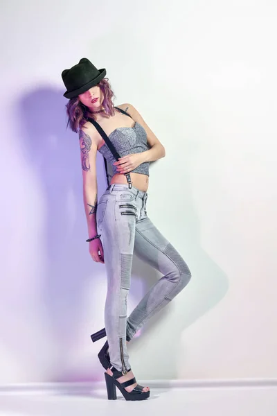 Chica sexy con el pelo morado y un tatuaje en el cuerpo posando sobre fondo gris. Mujer perfecta en jeans grises y una camiseta, maquillaje hermoso y brillante . — Foto de Stock