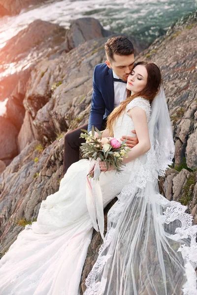 Belo casal apaixonado beijando enquanto sentado nas rochas perto do rio. Casamento casal ao pôr do sol e rios, amor e sentimentos ternos. Casal amoroso a descansar. Cerimônia de casamento ao ar livre. Casal perfeito . — Fotografia de Stock