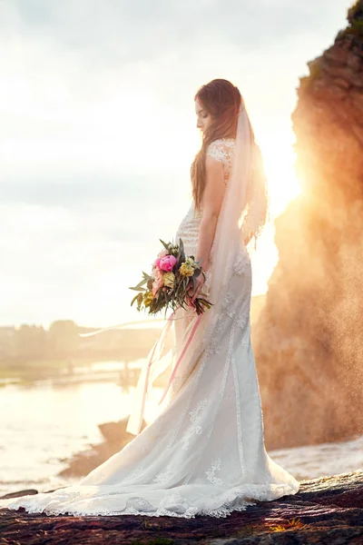 Bella donna sposa in piedi su rocce dal mare. Sposa al tramonto e surf, amore e sentimenti teneri. Donna amorevole che riposa. Cerimonia di matrimonio all'aperto. Matrimonio perfetto. Foto offuscata . — Foto Stock