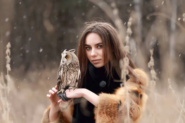 Frau im Pelzmantel mit Eule an der Hand beim ersten Herbstschnee. schöne Brünette mit langen Haaren in der Natur, in der Hand eine Eule. romantische, zarte Mädchen — Stockfoto