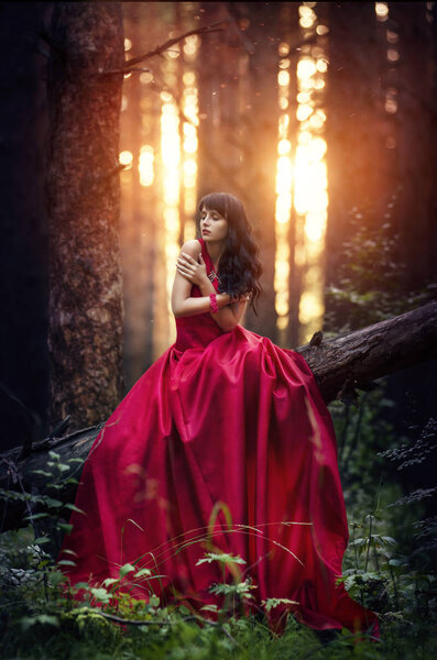 Woman in a long red dress alone in the forest. Fabulous and mysterious image of a girl in a dark forest in the evening sun. Sunset in the forest, the Princess got lost