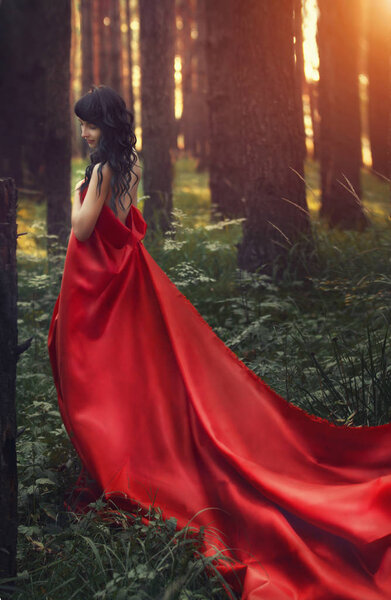 Woman in a long red dress alone in the forest. Fabulous and mysterious image of a girl in a dark forest in the evening sun. Sunset in the forest, the Princess got lost