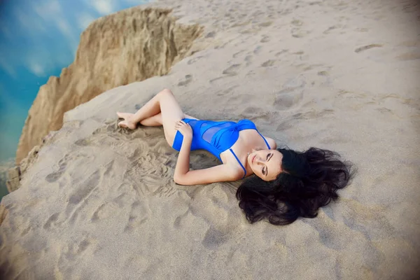 Жінка в блакитному купальнику на піску на пляжі біля моря лежить в променях вечірнього сонця. Красива жінка на тлі піщаних пагорбів . — стокове фото