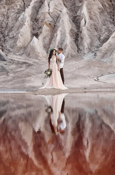 Ζευγάρι στην αγάπη αγκαλιάζει το φόντο βουνά και κόκκινη λίμνη. Τοπίο ρεαλιστικό παραμύθι, τα βουνά του Άρη. Γυναίκα με ένα μπουκέτο λουλούδια στα χέρια των ανδρών. — Φωτογραφία Αρχείου