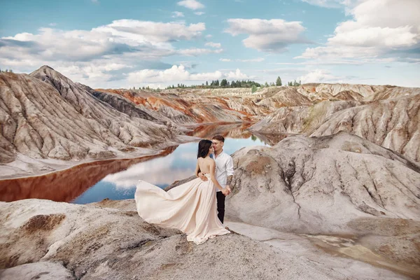 Ζευγάρι που στέκεται στο βάραθρο του βουνού και η κόκκινη λίμνη και την αγκαλιάζει. Η νύφη και ο γαμπρός στην αγάπη, η γυναίκα στο μακρύ φόρεμα στα χέρια των ανδρών. — Φωτογραφία Αρχείου