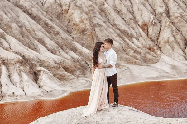 Casal fica no precipício da montanha e do lago vermelho e abraçando. A noiva e o noivo apaixonados, a mulher de vestido comprido nos braços dos homens . — Fotografia de Stock